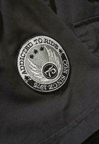 Črn obliž z napiso "Odvisen od vožnje", z obrobami in zvezdami, izvezen v srebrni barvi. Krožna zasnova z teksturiranim zaključkom.