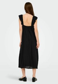Mujer con cabello largo castaño que lleva un vestido midi negro con tirantes de hombro rizados y una espalda baja descubierta, combinado con zapatos negros de tacón bajo.