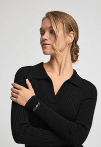 Schwarzer gerippter Pullover mit V-Ausschnitt und Polokragen. Hat taillierte Ärmel und gestickte Details am Bund. Glatte Textur.