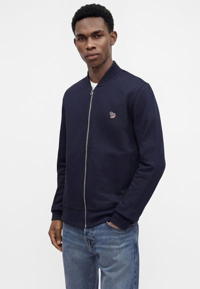 Homme portant une veste zippée bleu marine avec un petit logo zèbre multicolore sur la poitrine, jean bleu, debout avec une main dans la poche.
