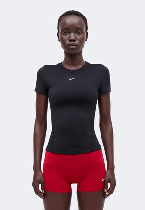 Femme portant un haut noir ajusté à manches courtes Nike et un short de sport rouge Nike, debout devant un fond clair uni.