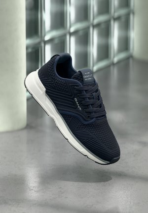 Baskets athlétiques bleu marine avec empeigne en mesh texturé, semelle blanche et accents foncés. Comprend des lacets et un marquage sur la languette et le talon.