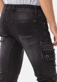 Jean en denim noir avec une coupe slim, poche cargo zippée et logo sur la poche arrière. Tissu texturé avec des coutures visibles.