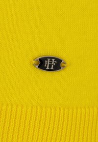 Tejido de punto en un amarillo brillante con una textura acanalada. Cuenta con una pequeña etiqueta ovalada de metal negro con acentos plateados y un logotipo.