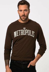 Maglietta a maniche lunghe marrone con grafiche "METROPOLIS" in bianco opaco, collo rotondo, polsini a costine e tessuto morbido. Jeans neri.