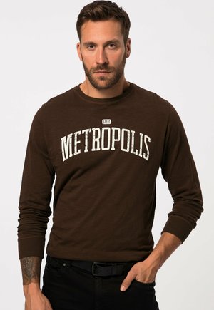 Ruskea pitkähihainen t-paita, jossa on kuluneen valkoinen "METROPOLIS" -grafiikka, pyöreä kaula-aukko, ribbiraidalliset hihansuut ja pehmeä tekstuuri. Mustat farkut.