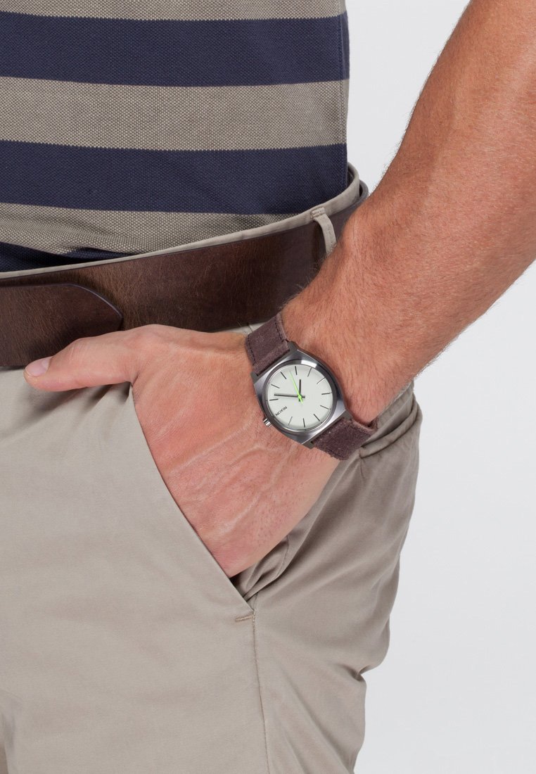 Mann mit braunem Leder-Gürtel, beigen Hosen und einem gestreiften navy-blauen und grauen Hemd, mit der linken Hand in der Tasche, die eine Uhr mit braunem Armband zeigt.