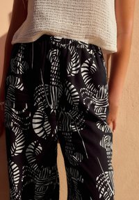 Pantalon noir avec un motif graphique de feuilles blanches, taille élastique, tissu léger et coupe décontractée. Haut texturé blanc cassé visible au-dessus.