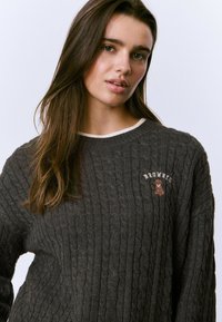 Suéter gris de punto con patrón de trenzas, que cuenta con un cuello redondo en color crema y un logo bordado de "Brownie" con un diseño de oso en el pecho.