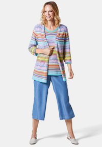 Kleurrijke, gestreepte cardigan in pastelkleuren over een effen blauwe top, gecombineerd met cropped blauwe denim broeken en lichte instapschoenen.
