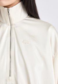 Kremo spalvos zip-up striukė su aukšta apykakle, kuri turi mažą siuvinėtą logotipą. Audinys yra sklandus ir minkštas.