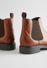 Brown leather Chelsea boots featuring elastic side panels, perforated accents, and rubber soles. The design is streamlined with a slightly raised heel.