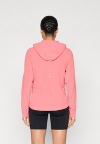 Ελαφρύ ροζ μπουφάν με κουκούλα και μακριά μανίκια, με μαλακή υφή και διακριτικές αντανακλαστικές λεπτομέρειες στην πλάτη. Συνδυάζεται με μαύρο σορτς.