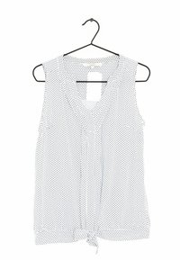 Blouse sans manches blanche à pois noirs, décolleté en V et détail de ourlet noué. Tissu léger, suspendue à un cintre noir.
