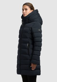 Mujer vestida con un abrigo acolchado largo y negro de invierno, con cuello alto y capucha, cerrado con cremallera, de pie de perfil contra un fondo liso.