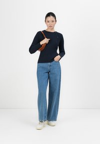 Junge Frau in einem dunkelblauen Rippenpullover, weiten blauen Jeans, weißen Sneakern und einer braunen Umhängetasche, die vor einem weißen Hintergrund steht.