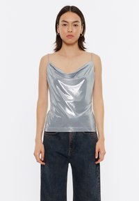 Top sin mangas de cuello en cowl de color plateado con tirantes delgados. Material brillante, diseño ajustado y textura suave. Combinado con jeans de pierna ancha oscuros.