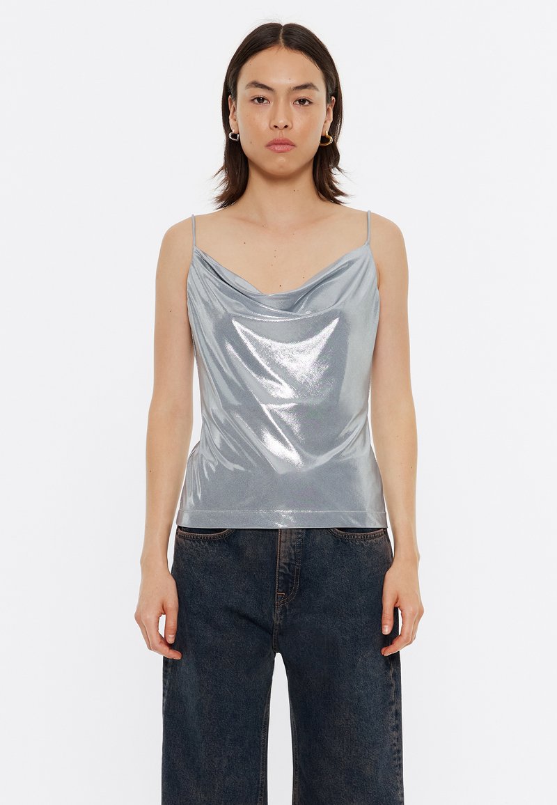 Top sin mangas de cuello en cowl de color plateado con tirantes delgados. Material brillante, diseño ajustado y textura suave. Combinado con jeans de pierna ancha oscuros.