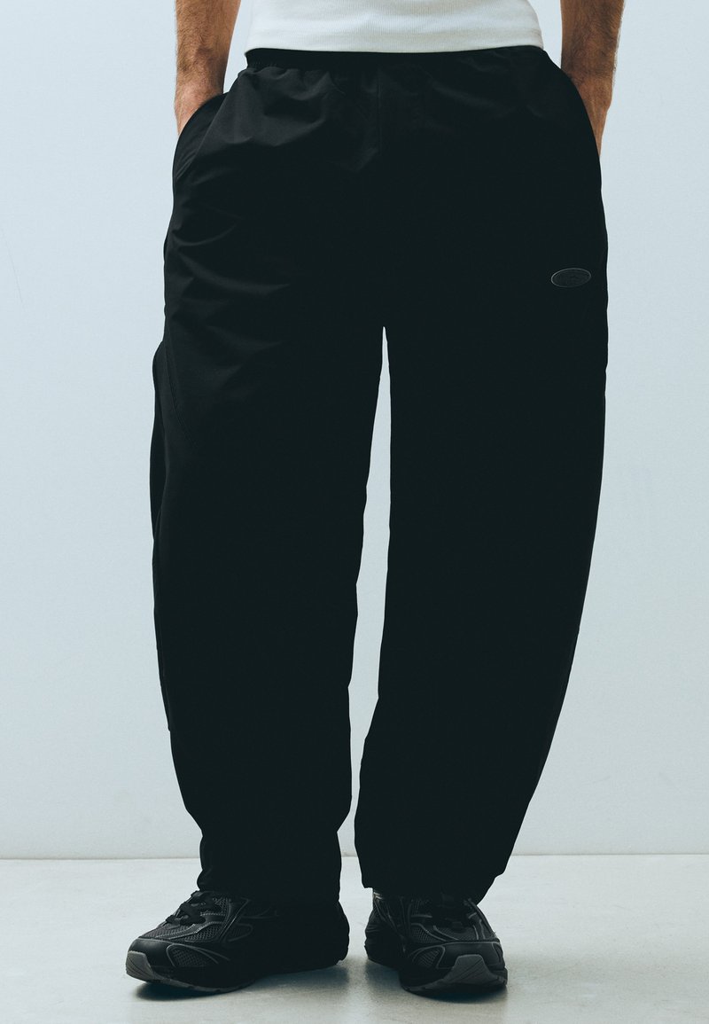 Homme portant un pantalon noir ample et des baskets noires, debout les mains dans les poches sur un fond uni.