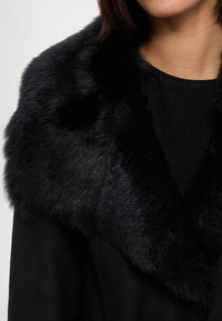 Fekete műszőrme gallér fekete kabáton, puha, textúrált felülettel és elegáns dizájnnal, amely kiemeli a meleget és a stílust.
