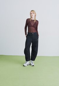 Blonde sieviete stāv ar rokām kabatās, ģērbta caurspīdīgā brūnā garo piedurkņu augšējā daļā, melnās plašās biksēs un baltās sporta apavos uz zaļā grīdas.