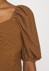 Brun puffärmad topp i texturerad tyg med mönstrade utskärningar, med fyrkantig halsringning och samlad detalj vid axeln.