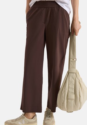 Personne portant un pantalon marron ample, des baskets beige, une chemise blanche ; tenant un grand sac matelassé beige par la sangle dans la main droite.