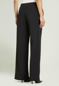 Pantalones anchos negros con textura suave, cintura elástica y diseño hasta el suelo, combinados con tacones altos negros.