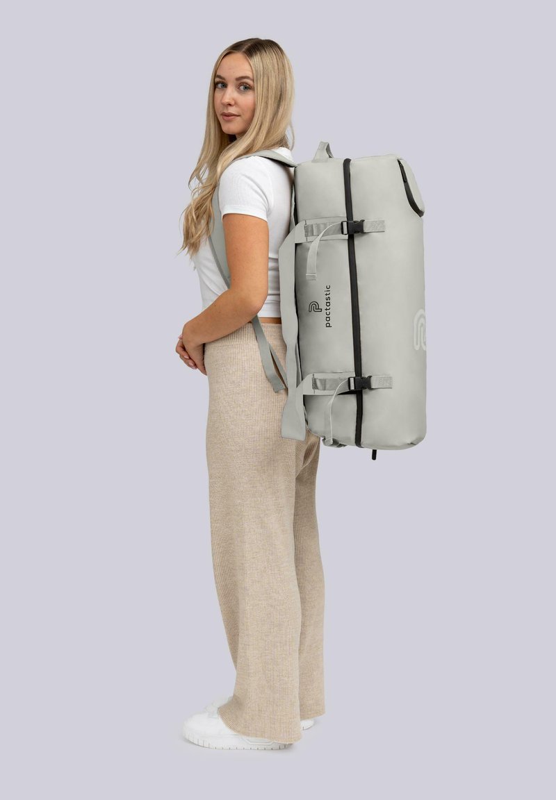Sac à dos gris en forme cylindrique, avec des bretelles ajustables et une fermeture éclair noire. Il présente un logo sur le côté et est fabriqué dans un matériau texturé et résistant.