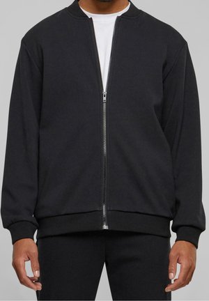Homme portant une veste noire zippée sur une chemise blanche, les mains détendues le long du corps sur un fond gris uni.