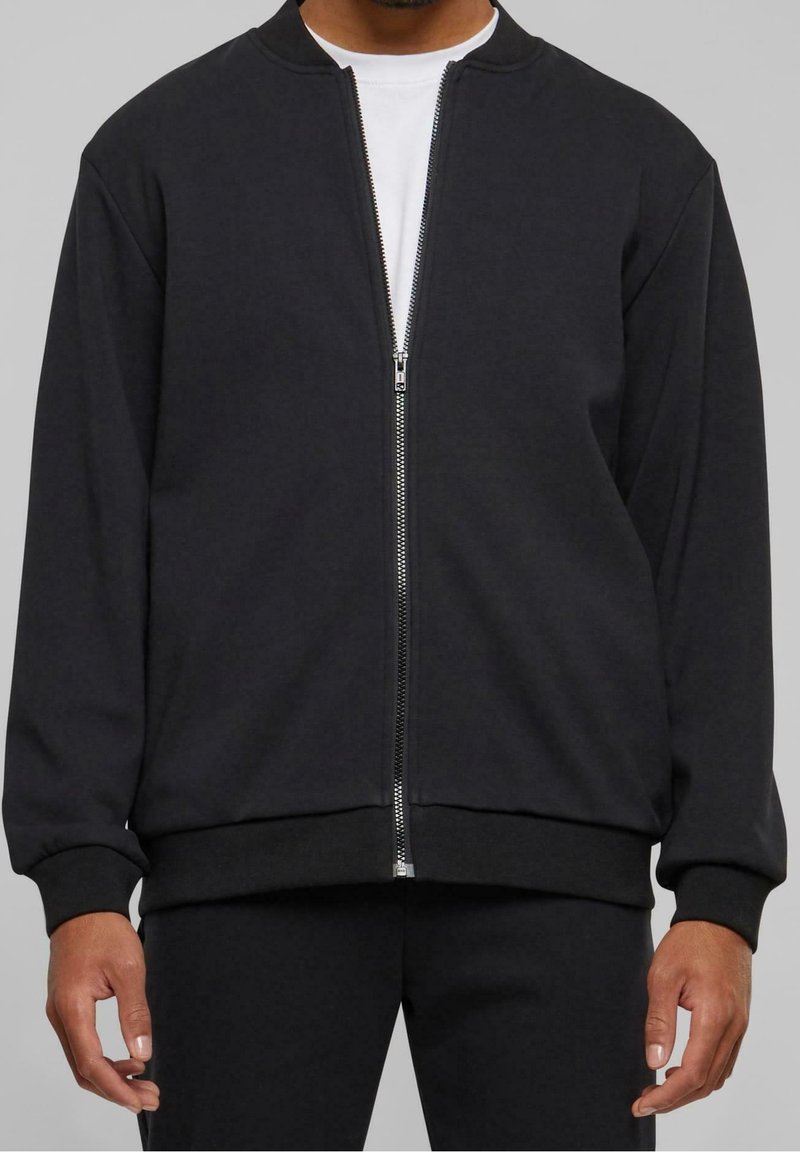 Homme portant une veste noire zippée sur une chemise blanche, les mains détendues le long du corps sur un fond gris uni.