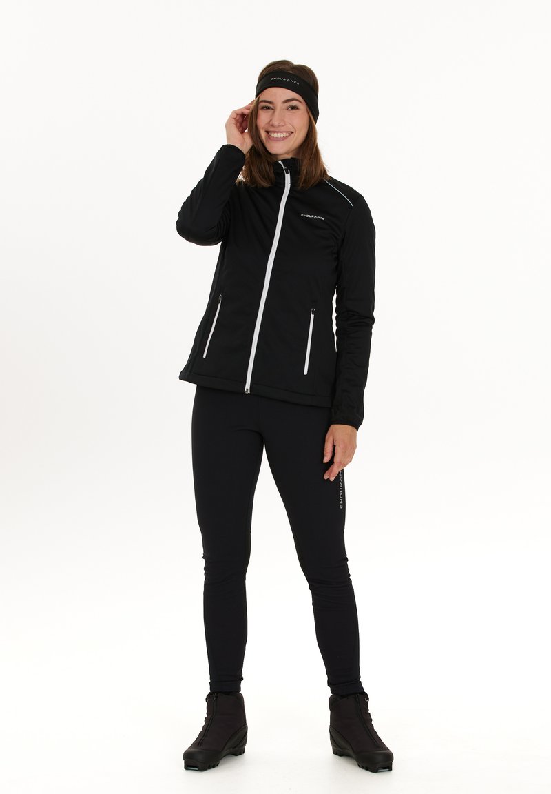 Musta urheilu takki, jossa on valkoinen vetoketju ja korostukset, yhdistetty mustiin legginsseihin ja saappaisiin. Mallilla on päässään yhteensopiva musta hiuspanta. Tasainen pinta.