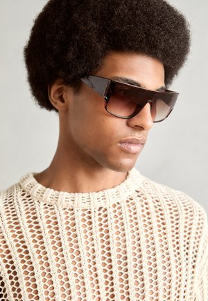 Sunglasses with a bold, angular design in dark tortoiseshell. Features tinted lenses and are worn with a loose, knitted cream top.