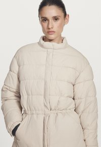 Donna con i capelli all'indietro, che indossa un piumino trapuntato beige con colletto alto e le mani nelle tasche, su uno sfondo neutro.