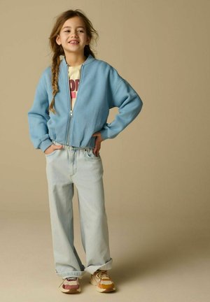 Fille souriante avec une longue tresse portant une veste à fermeture éclair bleue, un jean clair et des baskets colorées, se tenant devant un fond beige uni.