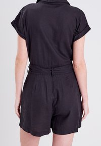 Rompers en lin noir avec des manches courtes, un col, une taille ceinturée et des shorts bien taillés, arborant un design épuré et minimaliste.
