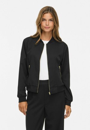 Femme portant une veste noire zippée sur une chemise blanche et un pantalon noir assorti, debout devant un fond clair uni.