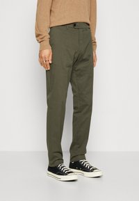 Olivgröna byxor i lättviktsmaterial, med midja med bältesöglor, sidofickor och raka ben, kombinerade med svarta sneakers.