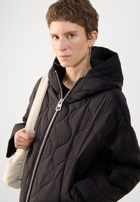 Veste en duvet noire matelassée avec capuche, dotée d'une fermeture éclair à l'avant et de poches latérales. Texture lisse et légère. Accentuée par une fermeture éclair argentée.