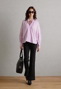 Helle rosa Bluse mit einer Schleifenkragen, schwarze ausgestellte Hose, schwarze Handtasche und Sonnenbrille. Glattes Material mit zusätzlicher Struktur an den Ärmeln.