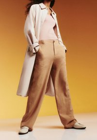 Beżowe lniane spodnie typu wide-leg o gładkiej fakturze, zestawione z jasnoróżową dopasowaną bluzką i kremowym długim płaszczem. Tło to gradient w odcieniach żółtego.