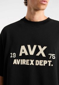 Schwarzes Baumwoll-T-Shirt mit kurzen Ärmeln, auf der Vorderseite mit beigem gesticktem Text "AVX 1975 AVIREX DEPT.". Rundhalsausschnitt.