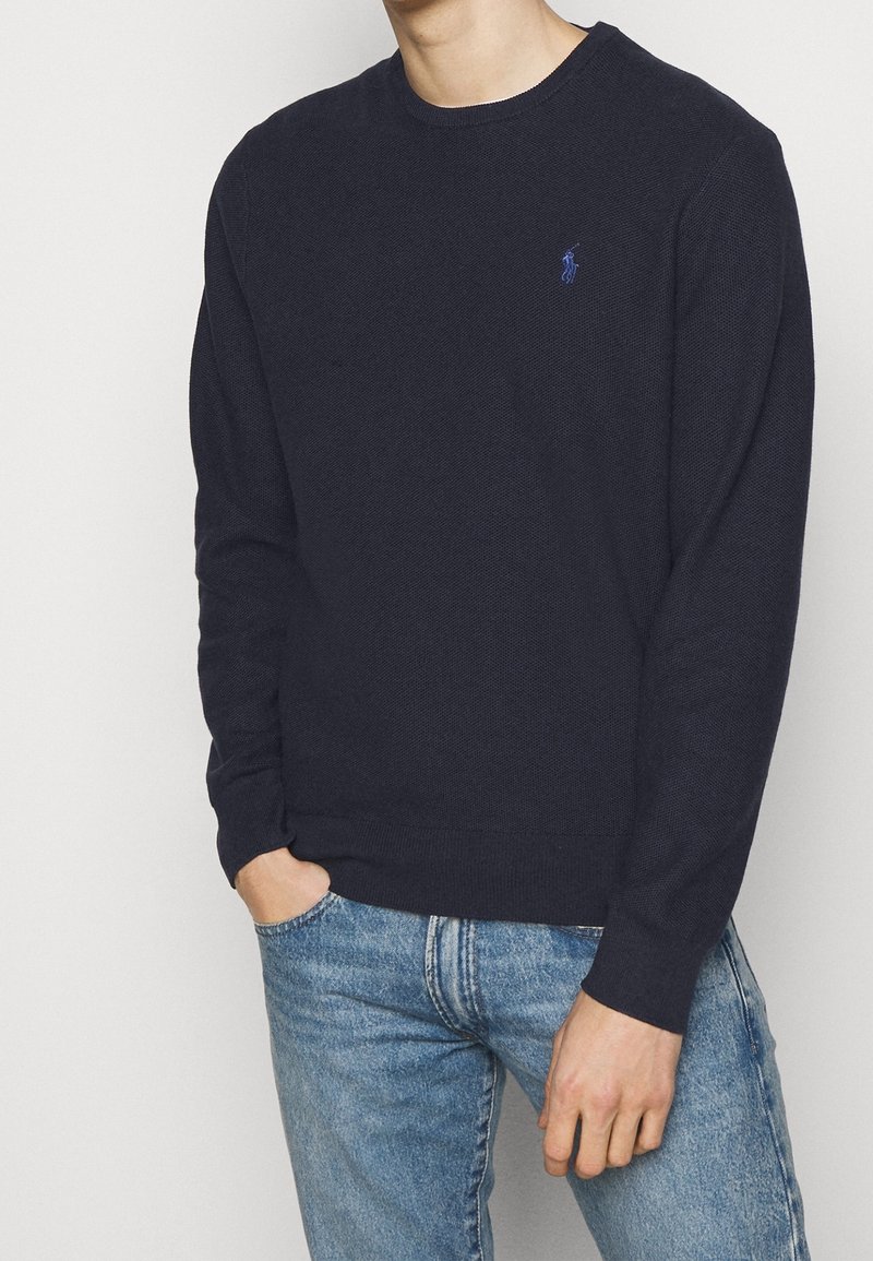 Homme portant un pull bleu marine à manches longues avec un petit logo brodé et un jean en denim bleu clair, main dans la poche.