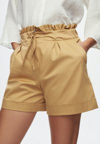 Tan shorts made of smooth fabric, featuring a ruffled elastic waistband, side pockets, and a folded hem for a relaxed fit.