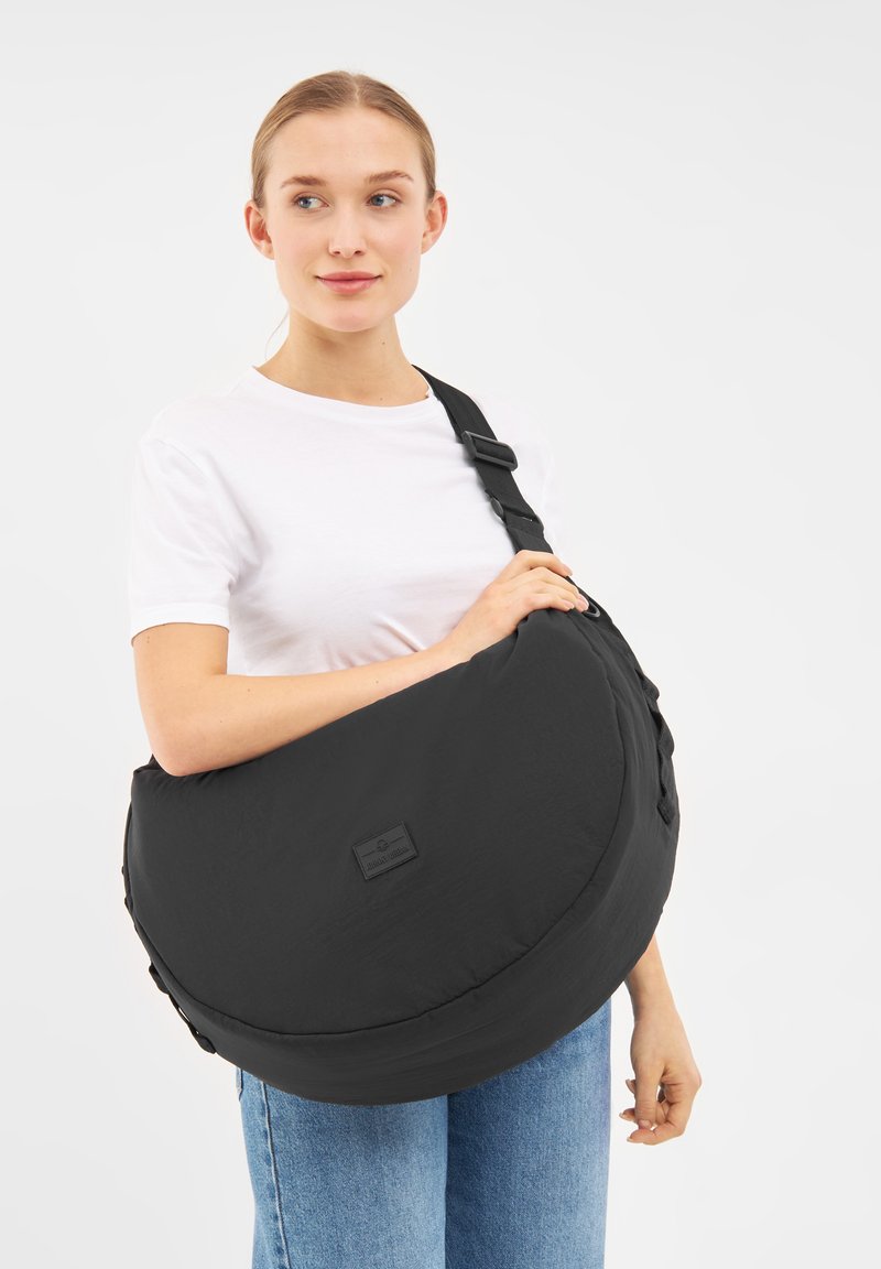 Černá přes shoulder taška vyrobená z měkké látky, s kulatým tvarem, nastavitelným popruhem a detailním logem na přední straně.