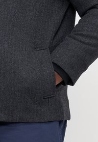 Dunkelgrauer Fischgratmantel mit strukturiertem Muster, ausgestattet mit einer Seiten- und schwarzen Manschettentasche. Weicher Stoff und maßgeschneiderter Schnitt.