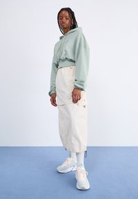 Sudadera corta de color verde claro con cordón; falda cargo crema con bolsillos; zapatillas en menta y blanco; modelo de pie sobre una alfombra azul.