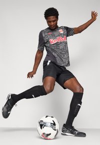 Maillot de football noir et gris avec le logo Red Bull, shorts noirs, chaussettes noires et crampons noirs ; joueur en position au-dessus d'un ballon de football noir et blanc.