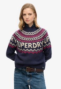 Maglione lavorato a maglia blu navy con collo alto, caratterizzato da motivi geometrici in crema, rosa e bordeaux, e la parola "SUPERDRY" in evidenza.