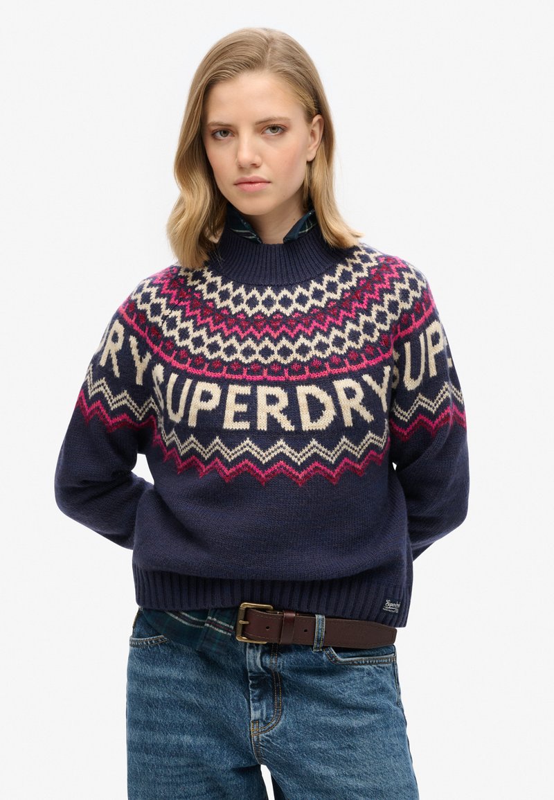 Maglione lavorato a maglia blu navy con collo alto, caratterizzato da motivi geometrici in crema, rosa e bordeaux, e la parola "SUPERDRY" in evidenza.